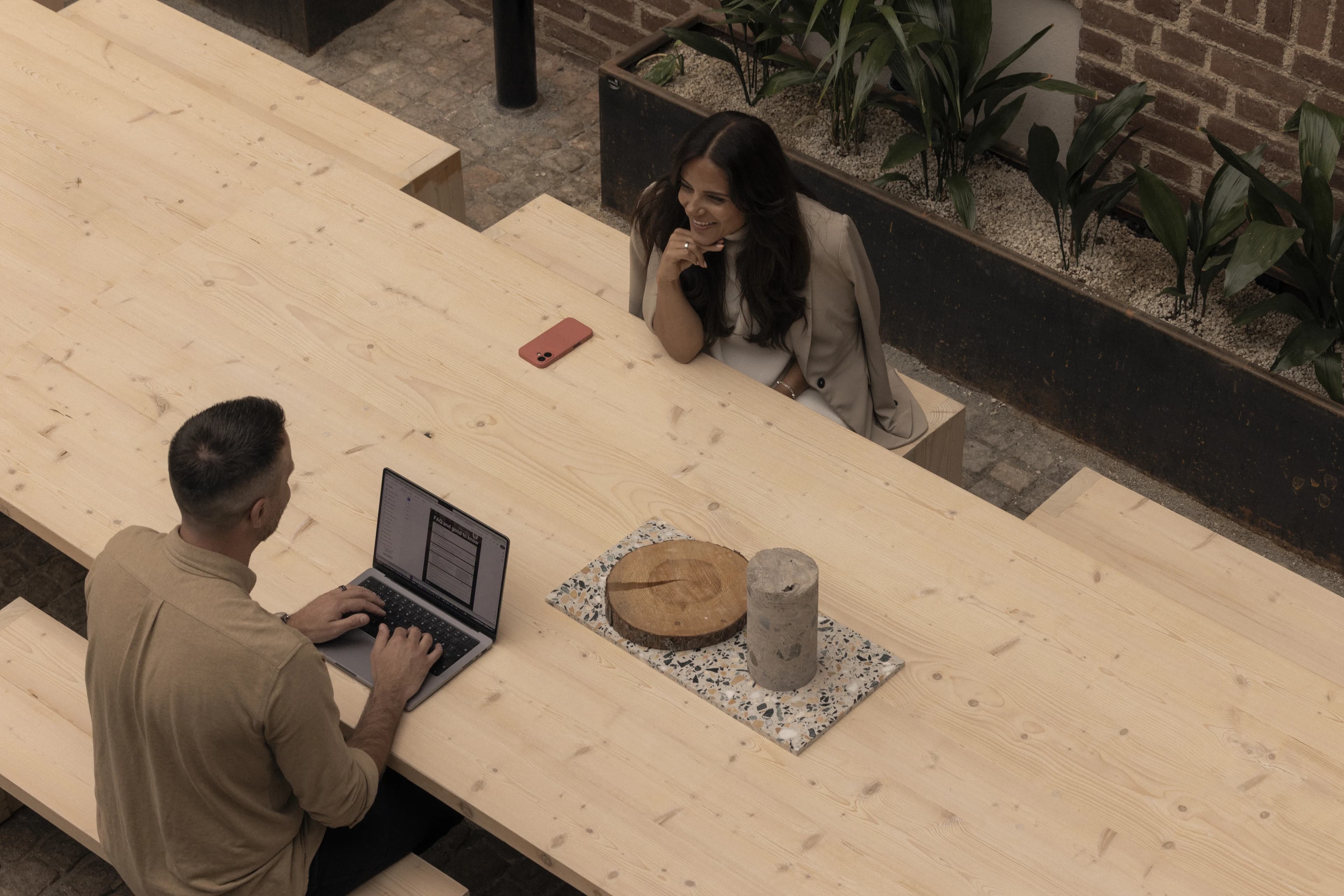 Woman in conversation with a man at a large wooden table, he has a laptop.